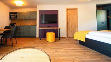 A compact room with a bed, a desk, a chair, a small stool, a television, a wardrobe, and a kitchenette.