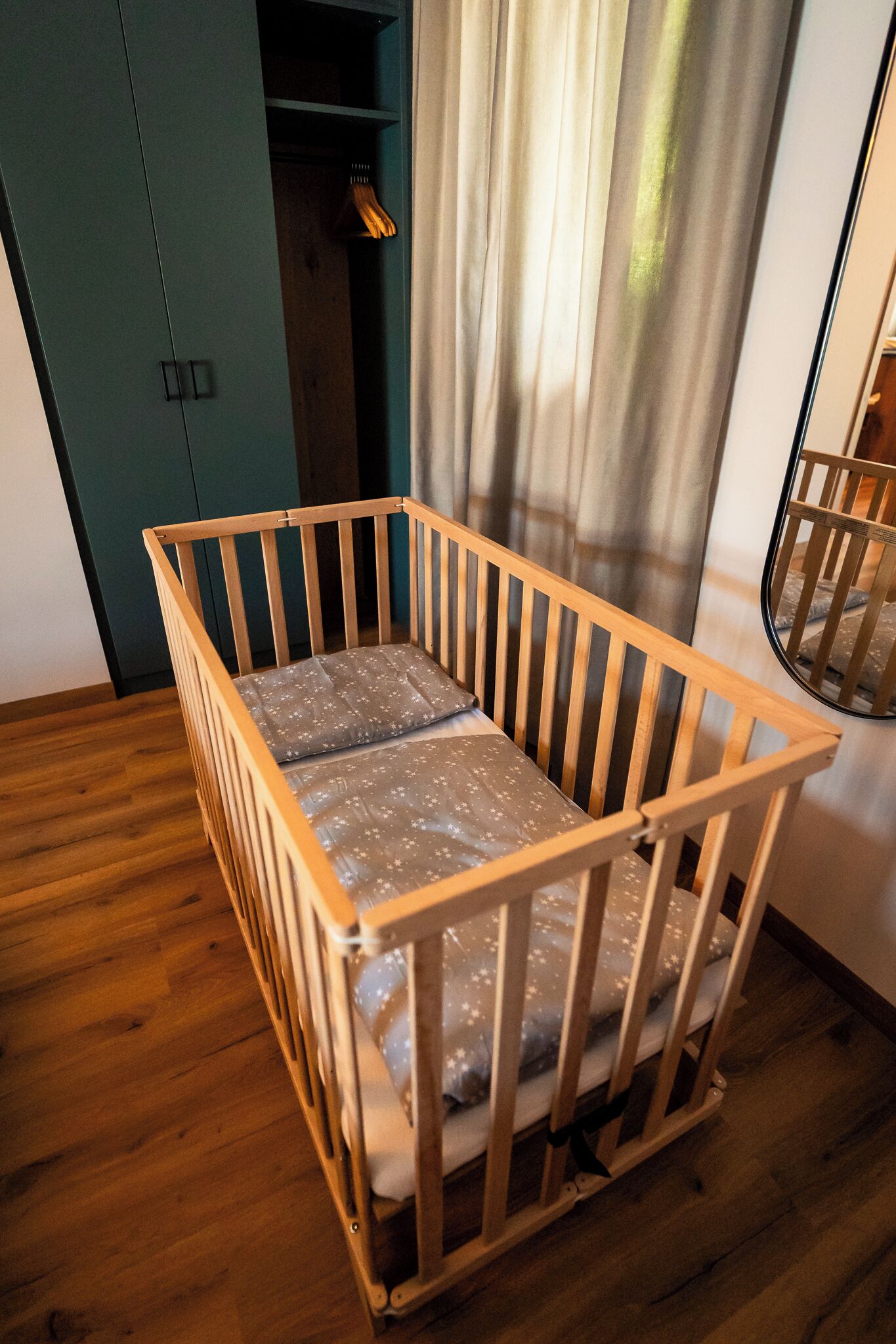 A wooden crib with a mattress, positioned in a room with wooden flooring and a green wardrobe in the background.