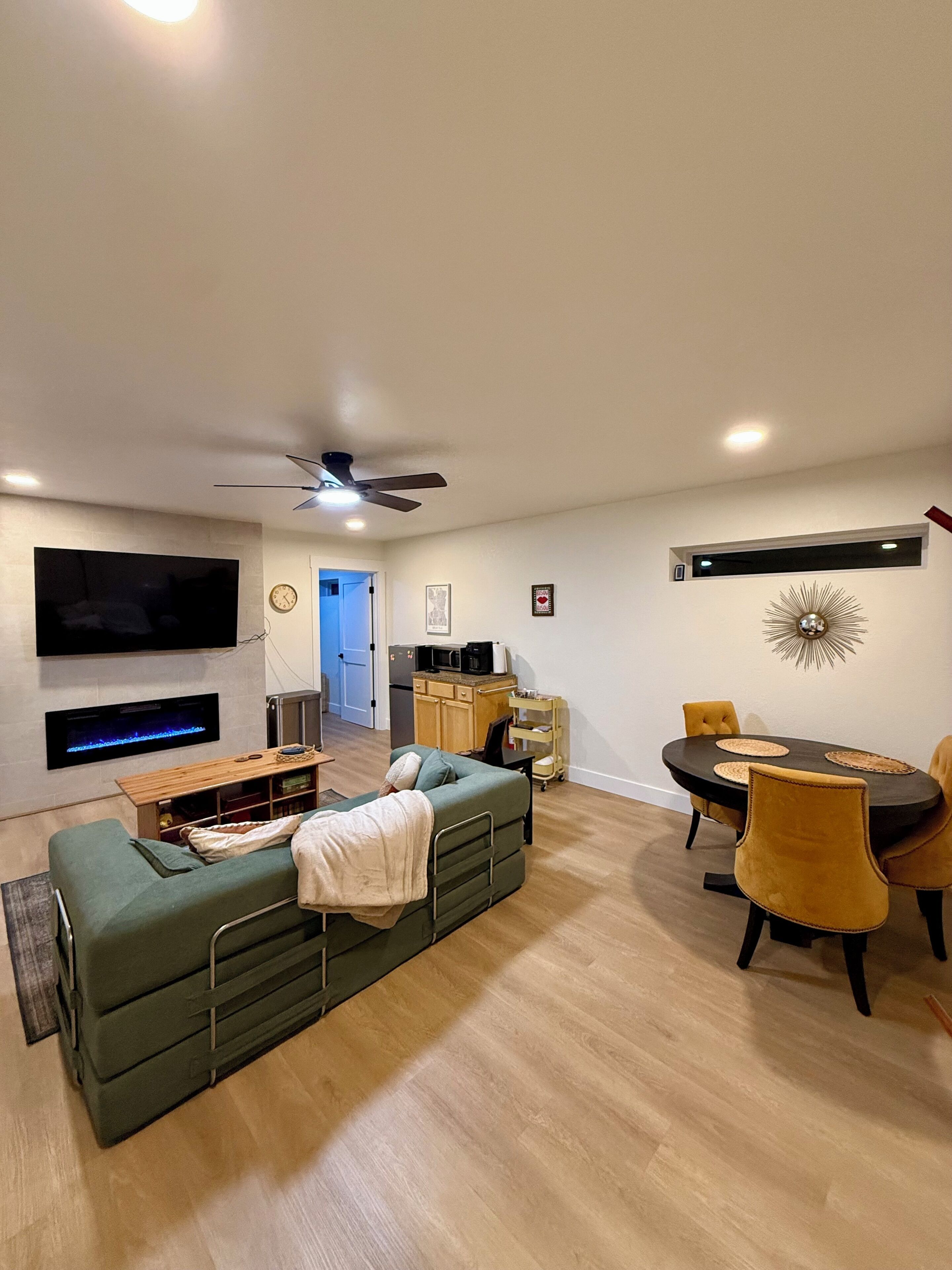 A modern living room with a flat-screen TV, a fireplace, a sofa, a dining table with chairs, and a ceiling fan.