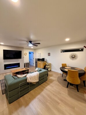 Ein modernes Wohnzimmer mit Fernseher, Kamin, einer Couch, einem Esstisch mit Stühlen und einem Deckenventilator.