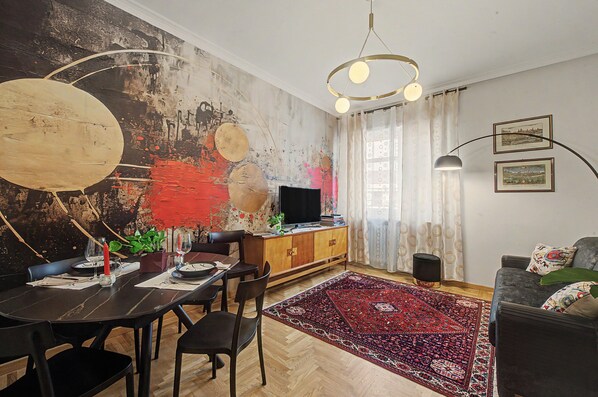 A modern living room with a dining area, a large abstract painting on the wall, a flat-screen TV, and a red patterned rug.