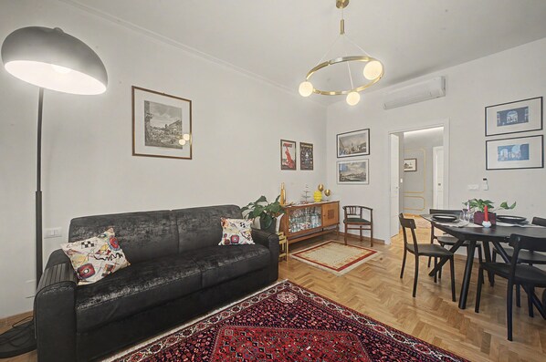 A modern living room with a black sofa, a dining table, and a red patterned rug.