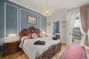 A bedroom with a large bed, ornate headboard, a chandelier, and a view of the city through a window.