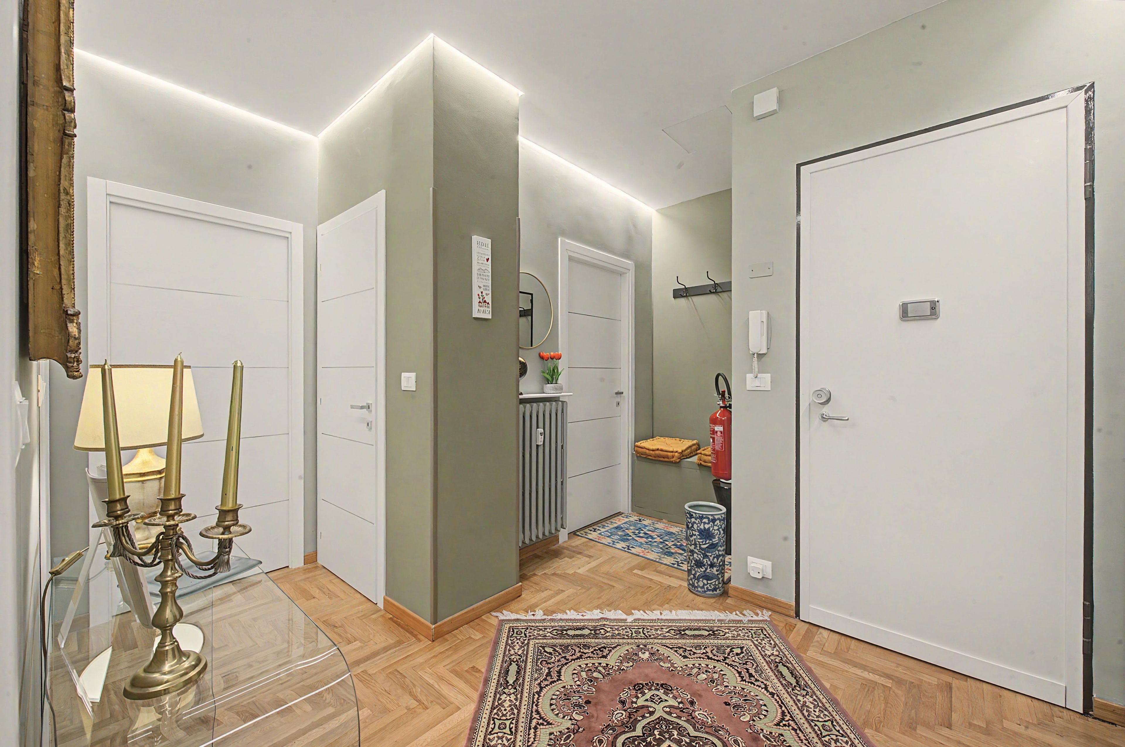 A hallway with a glass table, a candelabra, a rug, and a door with a fire extinguisher.