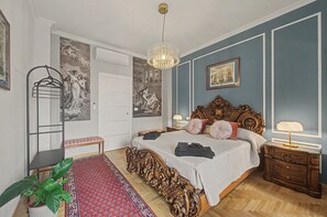 A bedroom with a large bed, ornate headboard, a hanging light fixture, a red patterned rug, and a potted plant.