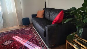 Ein modernes Wohnzimmer mit einem dunklen Sofa, einem roten Kissen, einer großen Grünpflanze, einem gemusterten Teppich und einem gerahmten Bild an der Wand.