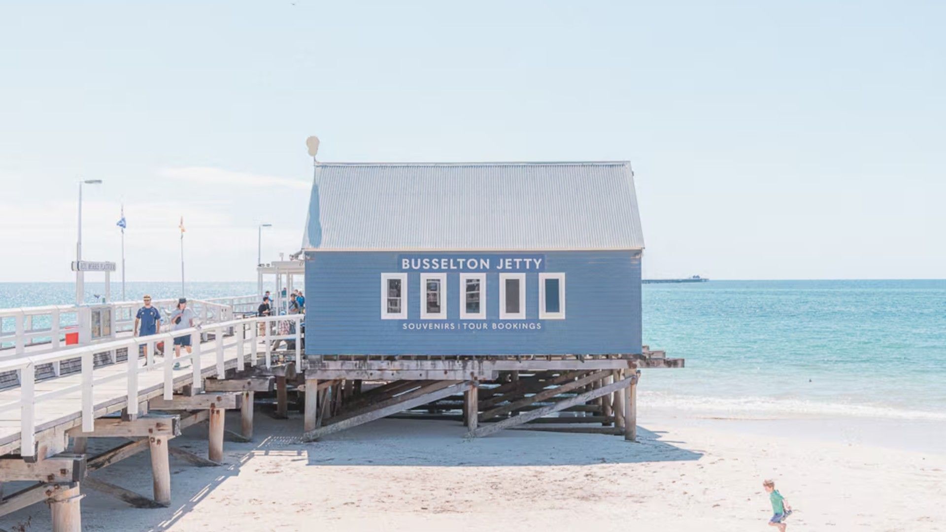 Un edificio blu con la segnaletica 'BUSSELTON JETTY', un molo che si protende nell'acqua e una spiaggia sabbiosa sullo sfondo.