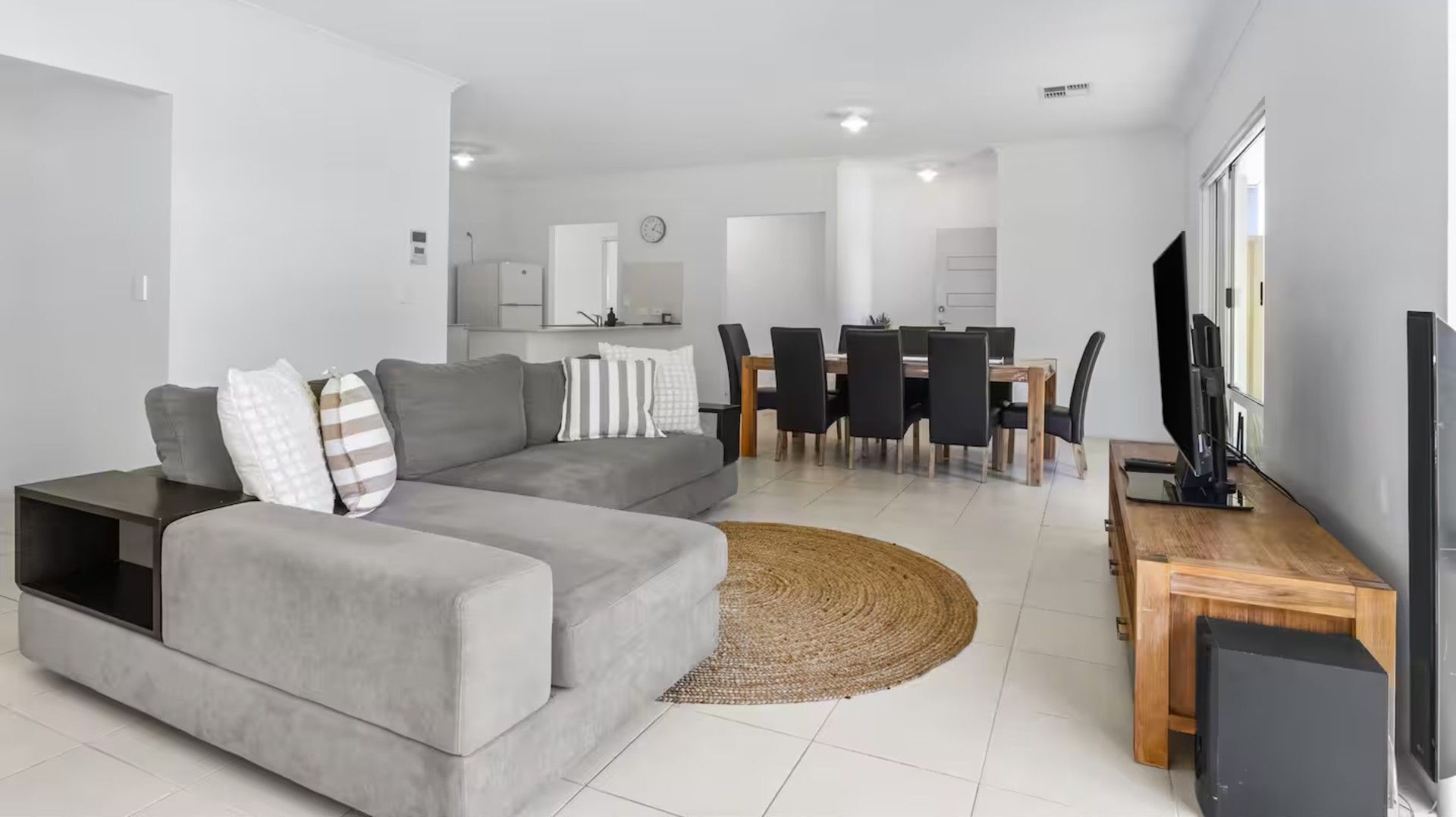 Un salon moderne avec un canapé gris, un coin repas doté d’une table et de chaises en bois, et un téléviseur à écran plat.