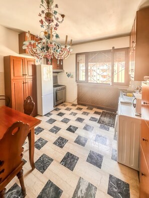 Une cuisine avec un sol carrelé, des meubles de rangement en bois, un réfrigérateur, une cuisinière et une fenêtre donnant sur des immeubles.