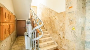 Un escalier en marbre avec une rampe métallique, des casiers en bois sur la gauche et un panneau d’avertissement sur le mur.