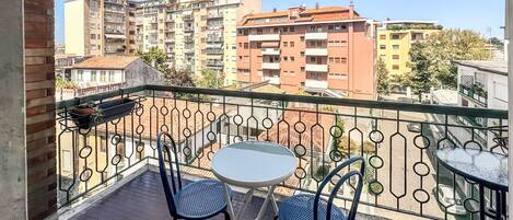 Een balkon met een ronde tafel en twee stoelen, met uitzicht op een stadslandschap met hoge gebouwen.