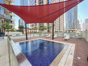 Una piscina en la azotea con un toldo rojo, rodeada de edificios modernos y sillones de descanso.