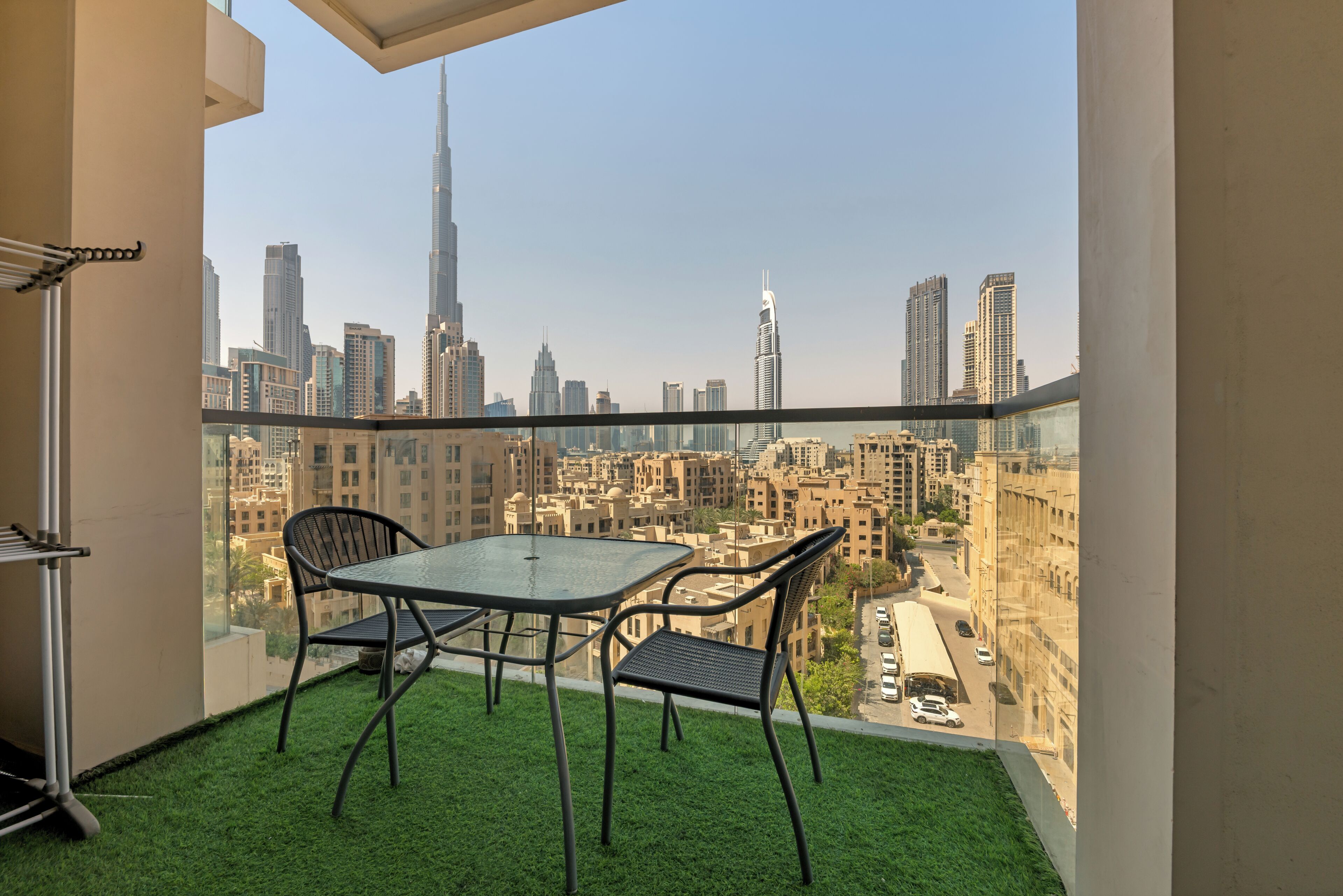 A balcony with a glass table and chairs, a small patch of artificial grass, and a cityscape view including tall buildings.