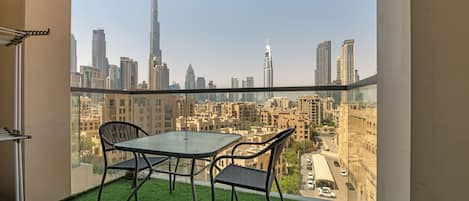 A balcony with a glass table and chairs, a small patch of artificial grass, and a cityscape view including tall buildings.