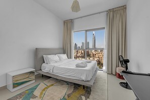 A modern hotel room with a bed, a desk, a chair, a standing fan, and a large window with a cityscape view. - Spacious 2Bedroom+ Maid room with Burj Khalifa View (دبي)