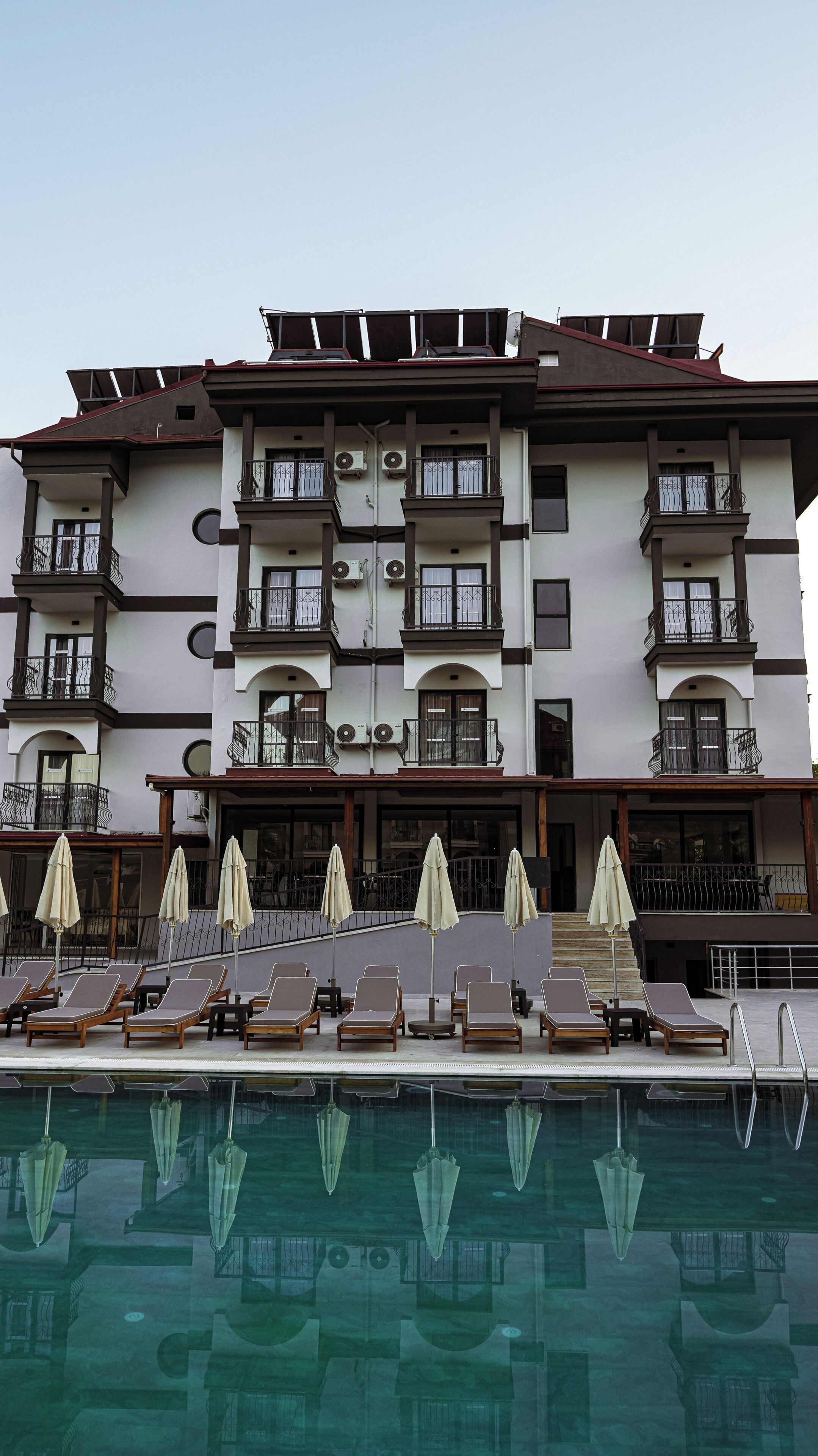 Un hotel de varios pisos con balcones, unidades de aire acondicionado y una zona de piscina con tumbonas y sombrillas.