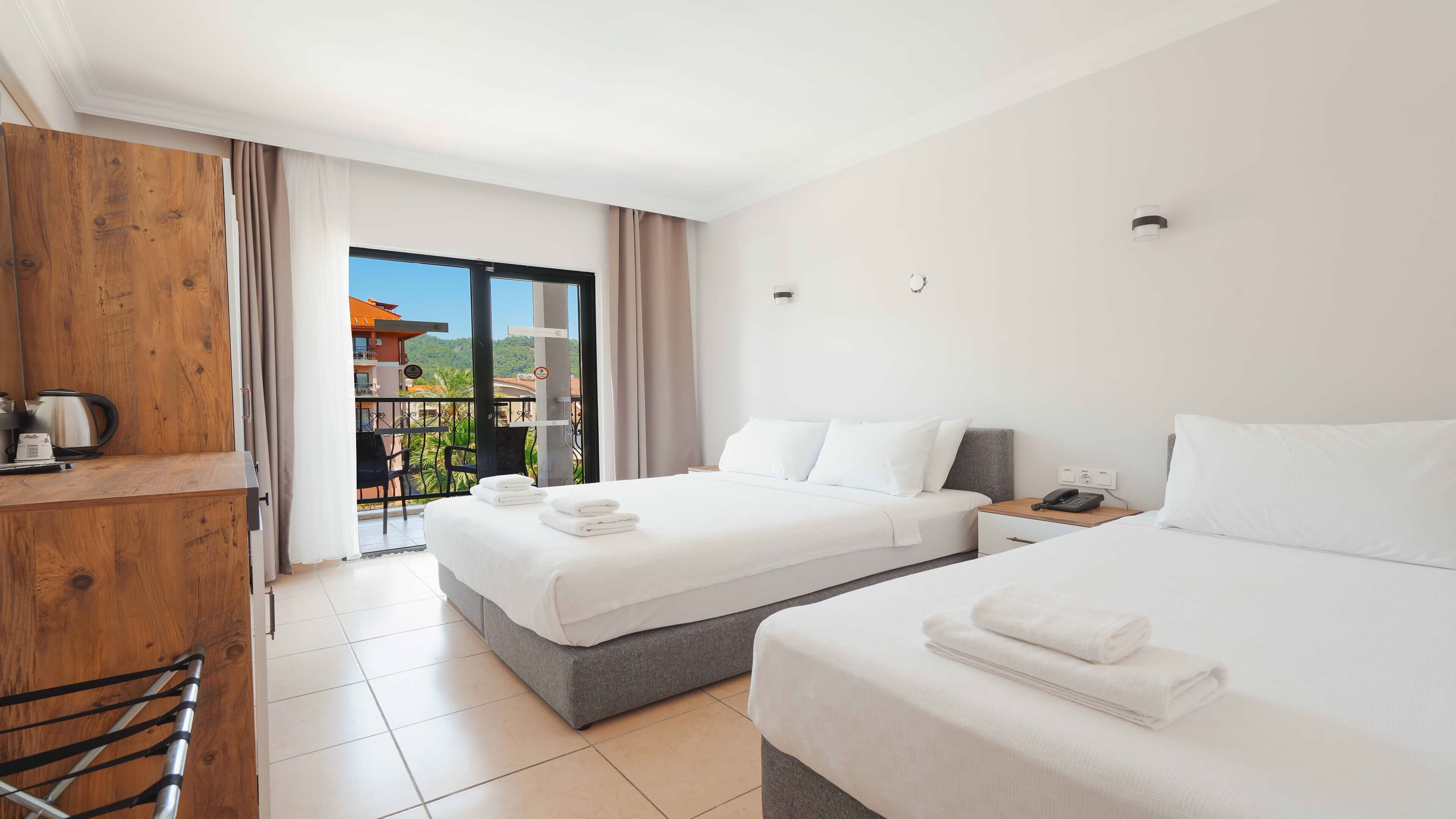 Habitación de hotel con dos camas, un escritorio con hervidor y un balcón con vistas a edificios y follaje.