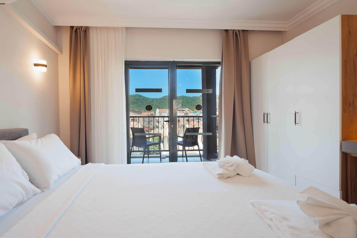 A hotel room with a bed, a balcony with a view of buildings and hills, and a wardrobe.