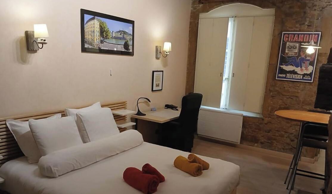 A hotel room with a bed, a desk, a chair, a window, and a wall-mounted lamp.