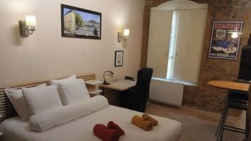 A hotel room with a bed, a desk, a chair, a window, and a wall-mounted lamp.