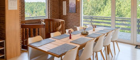 A dining area with a long table, chairs, and a view of a forest through large windows.