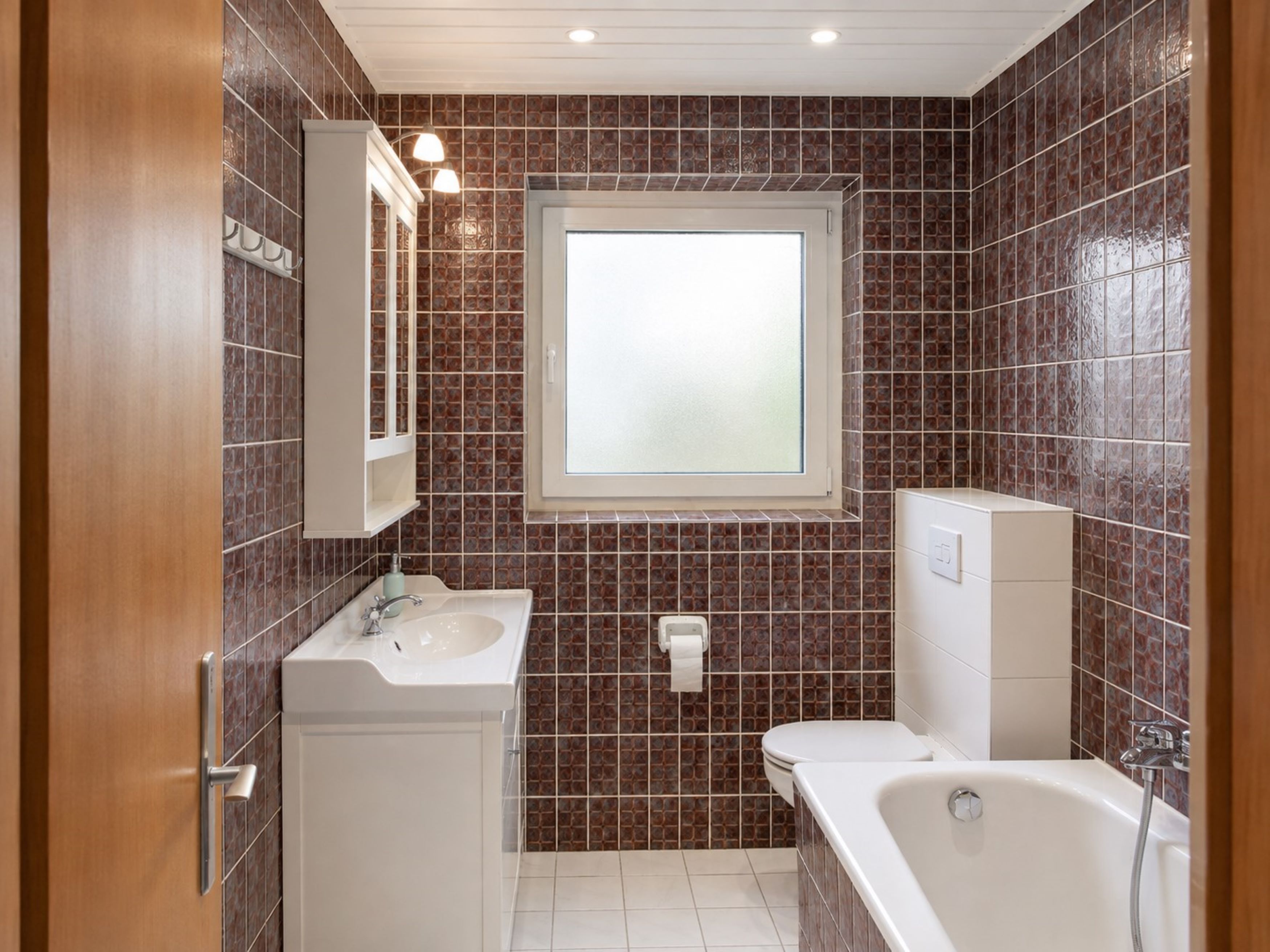 Ein Badezimmer mit einem weißen Waschbecken, einer Toilette und einer Badewanne. Über der Toilette befindet sich ein Fenster und über dem Waschbecken ein Spiegel.