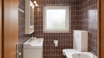Ein Badezimmer mit einem weißen Waschbecken, einer Toilette und einer Badewanne. Über der Toilette befindet sich ein Fenster und über dem Waschbecken ein Spiegel.