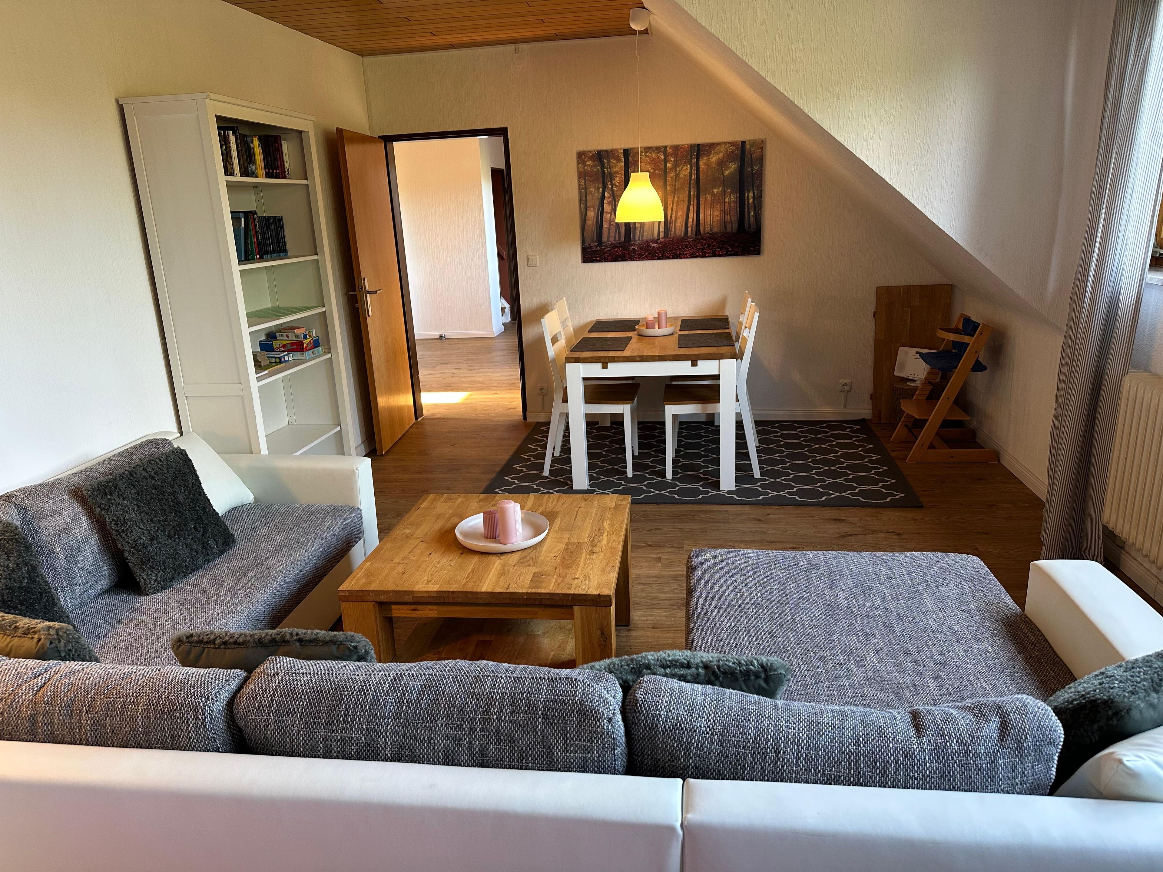 A cozy living room with a sofa, a coffee table, and a dining table with chairs. There is a bookshelf and a painting on the wall.