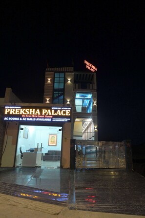 Un hotel llamado 'Preksha Palace' con fachada moderna, entrada visible y un letrero que indica disponibilidad de habitaciones con aire acondicionado y salones.