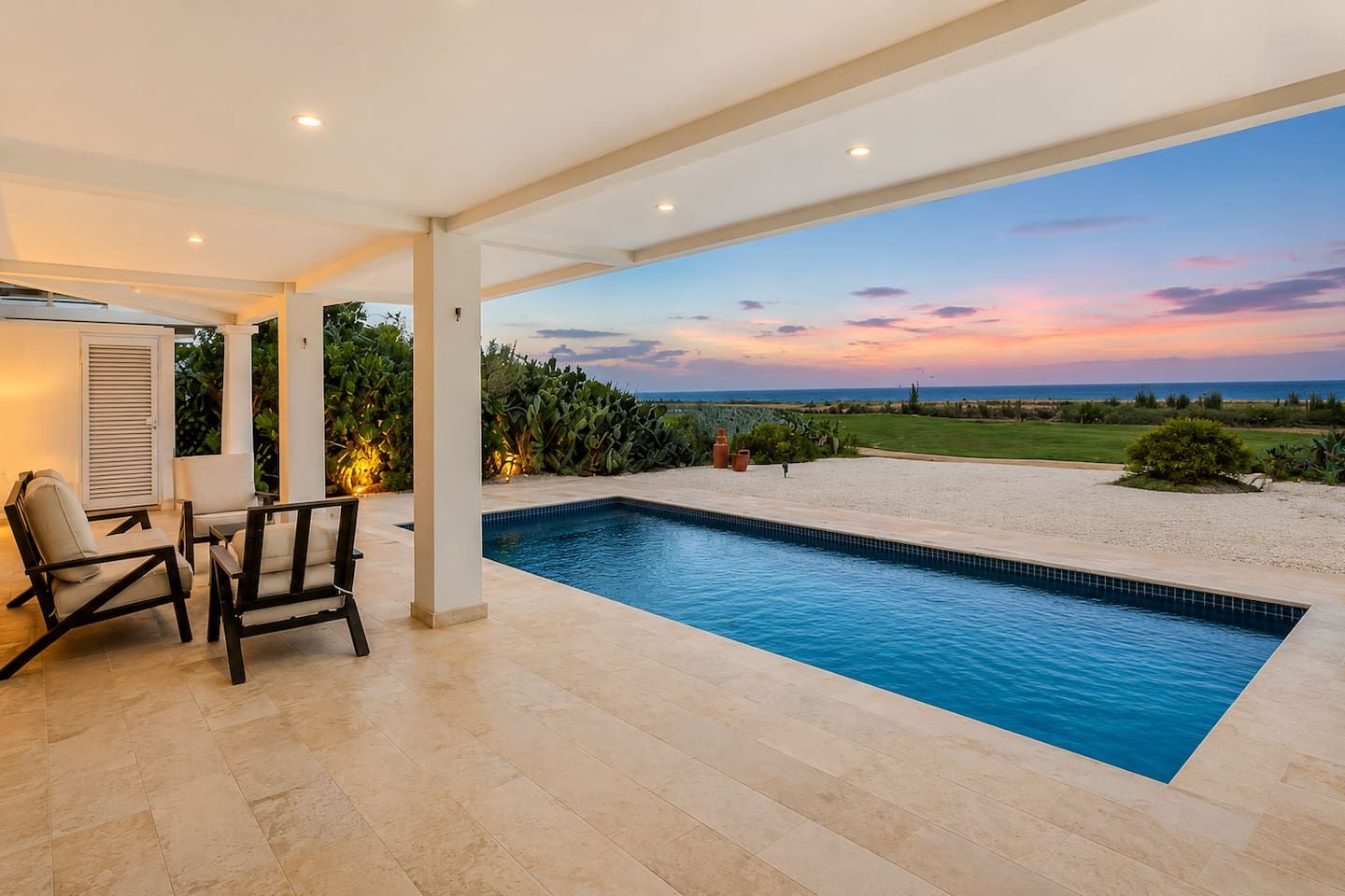 Una piscina con vista al océano al atardecer, un área de patio con sillas y una sección cubierta con una puerta.