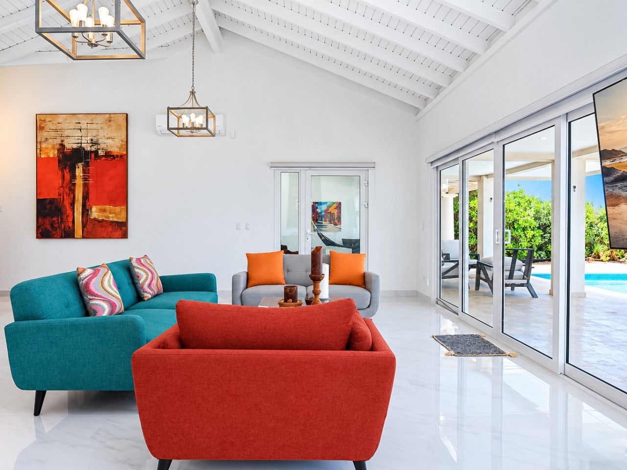 Un moderno salón con un sofá azul verdoso, un sillón rojo y un sofá gris con cojines naranja. Hay un cuadro abstracto grande en la pared, un candelabro y vistas a una piscina a través de puertas de cristal amplias.