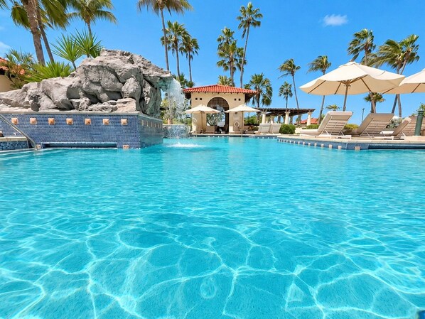 A clear blue pool with a rock formation and palm trees in the background.