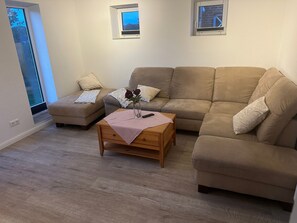 Un salon avec un canapé beige, une table basse en bois et une petite table d’appoint. Il y a deux fenêtres donnant sur l’extérieur.