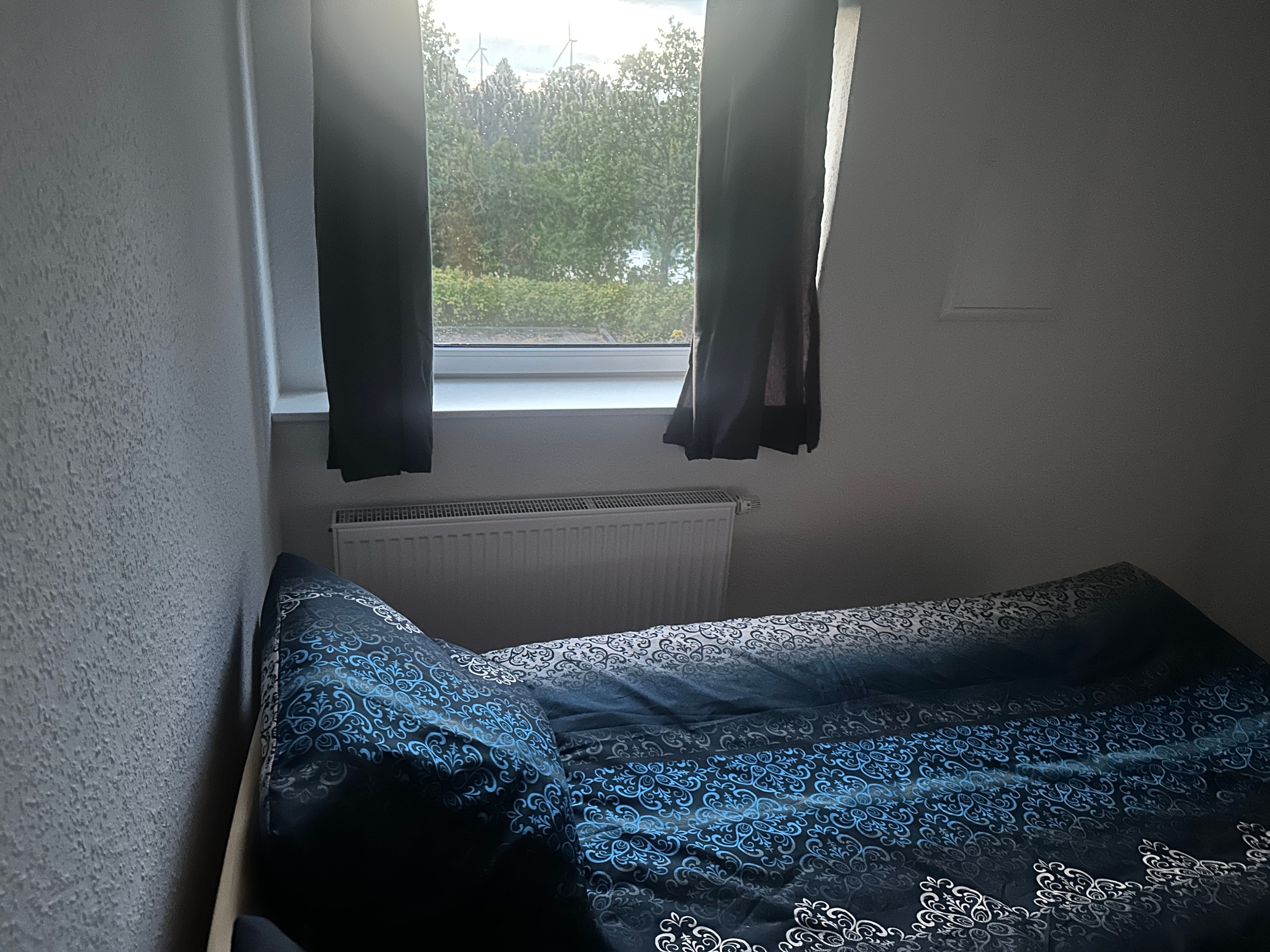 Ein kleines Zimmer mit einem Einzelbett, einem Fenster mit Vorhängen und einer Heizung.
