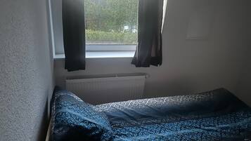 Ein kleines Zimmer mit einem Einzelbett, einem Fenster mit Vorhängen und einer Heizung.