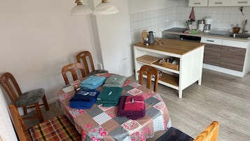 Een kleine keuken met een eethoek, houten stoelen, een tafel met een sprei en een hanglamp.