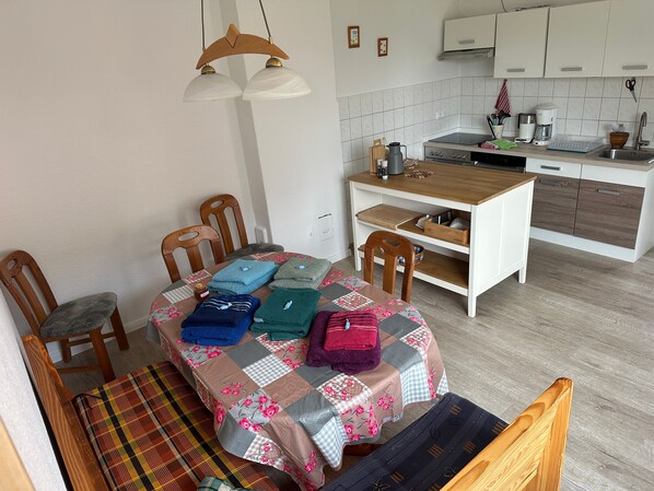 A compact kitchen with a dining area, wooden chairs, a table with a quilt, and a hanging light fixture.