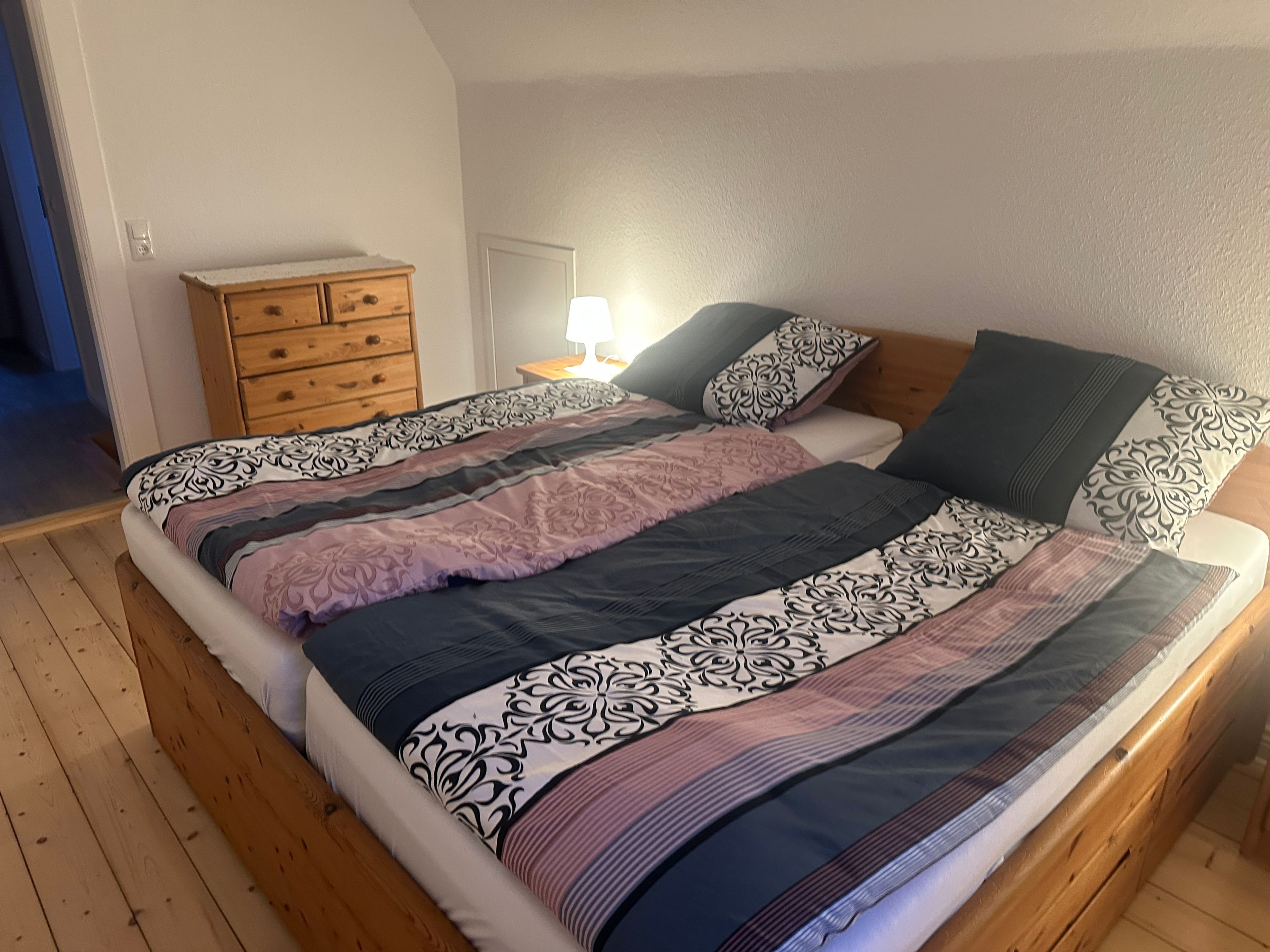 Een slaapkamer met een houten vloer, een bed met beddengoed met een patroon, een nachtkastje met een lamp en een kledingkast met laden.