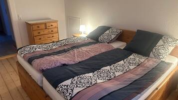 Een slaapkamer met een houten vloer, een bed met beddengoed met een patroon, een nachtkastje met een lamp en een kledingkast met laden.