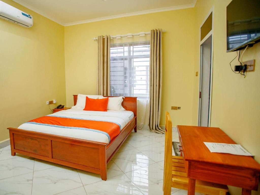 Habitación de hotel con cama, una mesa de madera, una silla, un televisor montado en la pared y una ventana con cortinas.