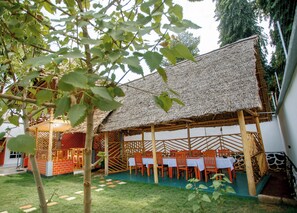Un área de comedor al aire libre con techo de paja, sillas y mesas de madera dispuestas para los huéspedes.