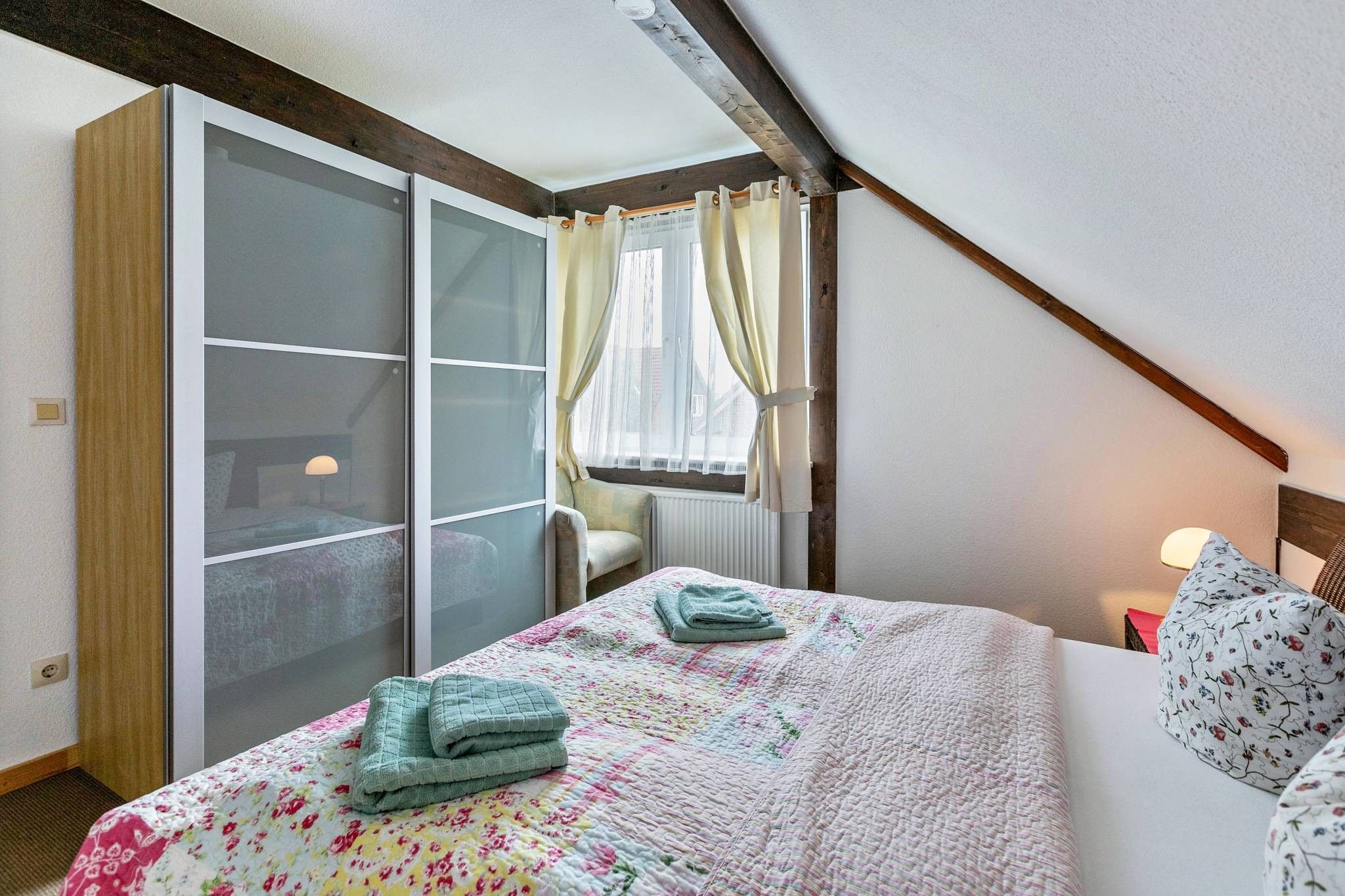 A bedroom with a bed, a window with curtains, a radiator, and a wooden ceiling beam.