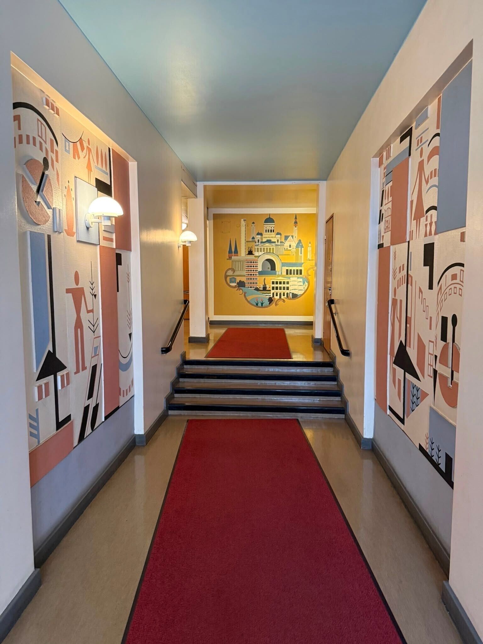 Un pasillo con alfombra roja que conduce a una escalera, paredes decoradas con arte abstracto y una lámpara en el techo.
