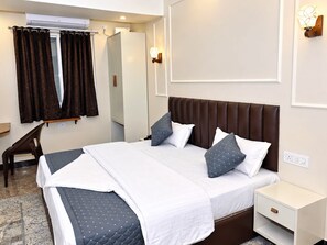 A hotel room with a large bed, a bedside table, a chair, an air conditioning unit, a wall-mounted light fixture, and a door. - Surya Stays (Prayagraj)