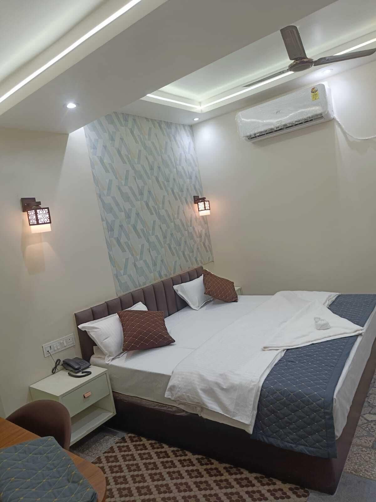 A hotel room with a bed, bedside table, lamp, and an air conditioning unit.