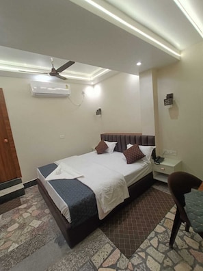 Una habitación de hotel con una cama, una mesita de noche, una silla, un equipo de aire acondicionado y un ventilador de techo.