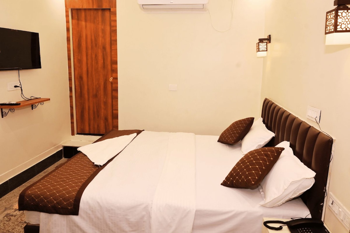 A hotel room with a bed, a TV mounted on the wall, an air conditioning unit, and a bedside telephone.