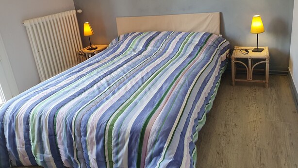 Una cama con colcha a rayas, dos lámparas de noche, una mesita de noche de madera y un radiador.