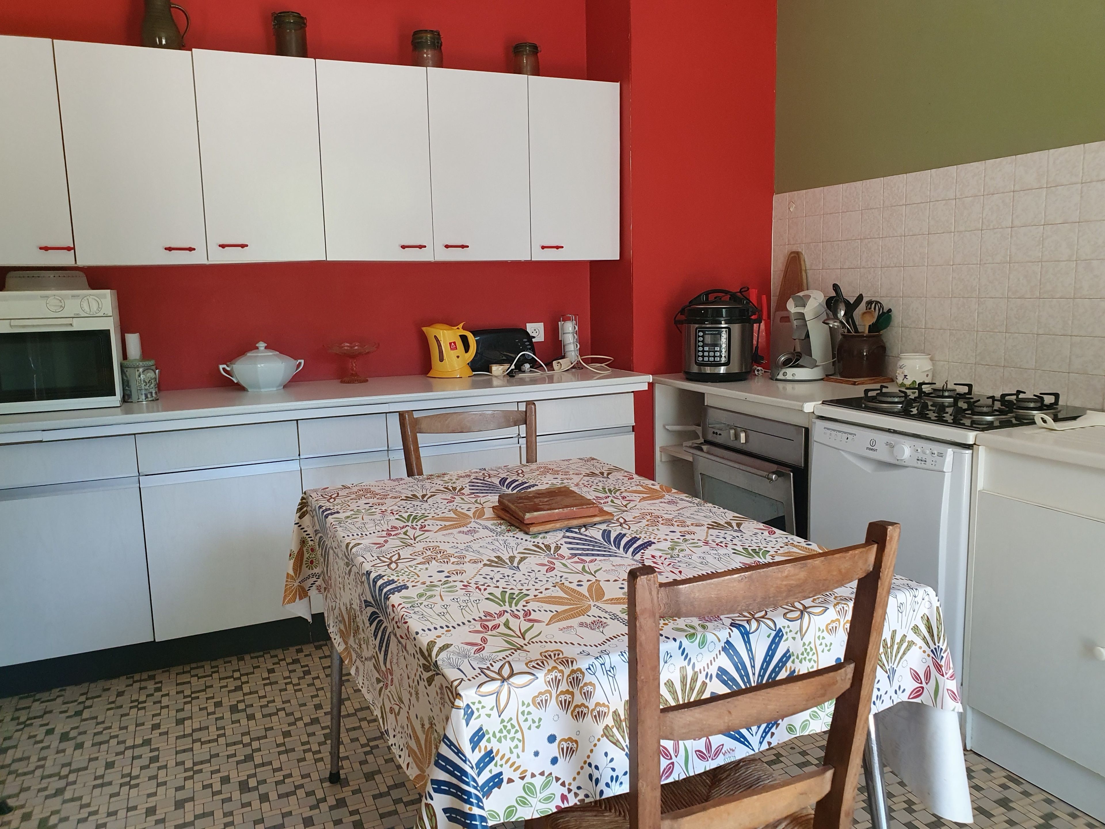 Een keuken met witte kastjes, een magnetron, een waterkoker, een fornuis en een tafel met een bloemendecoreerde tafelkleed.
