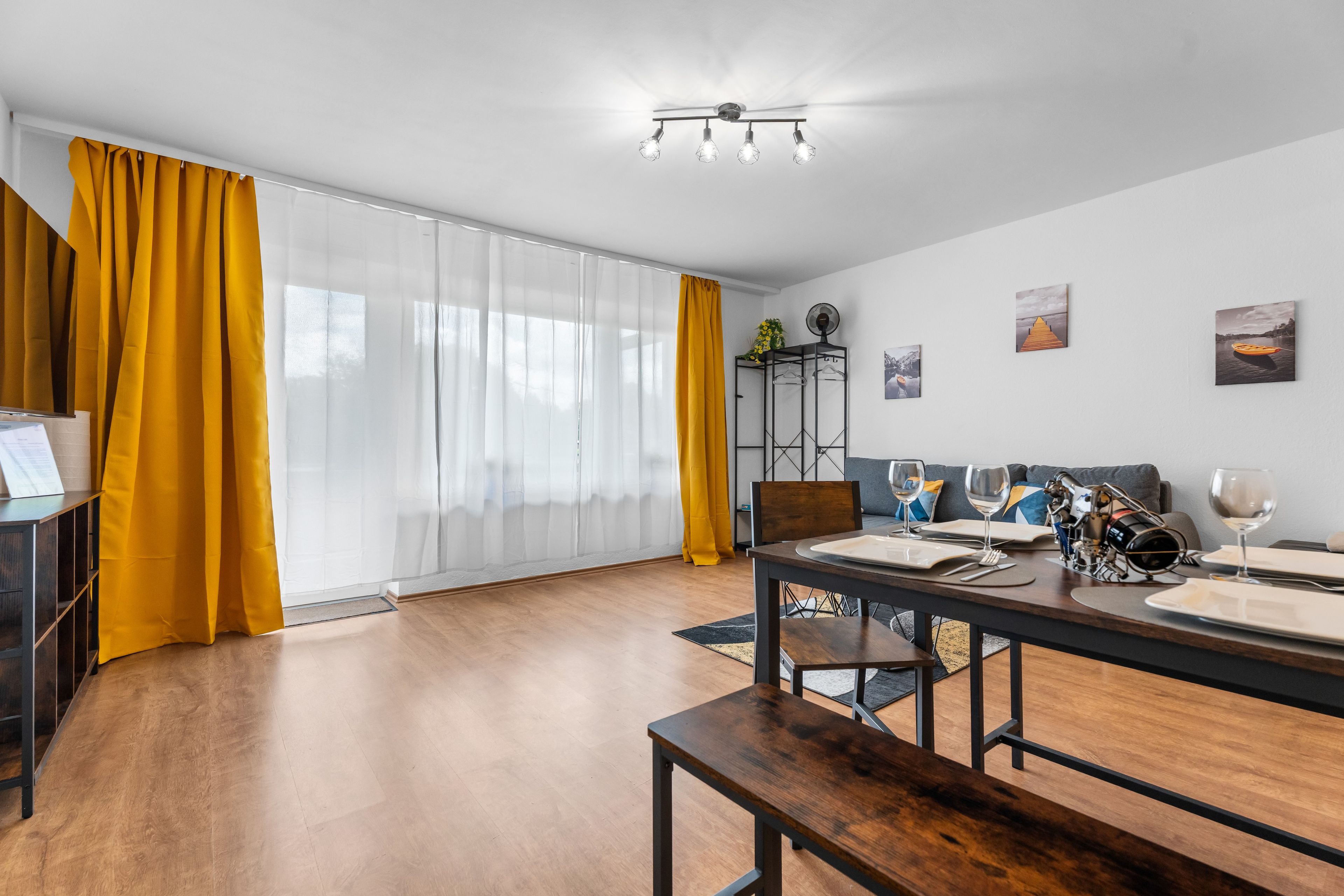 Un salon moderne avec un coin repas, comprenant une table en bois dressée pour deux personnes, un canapé et une grande fenêtre ornée de rideaux jaunes.